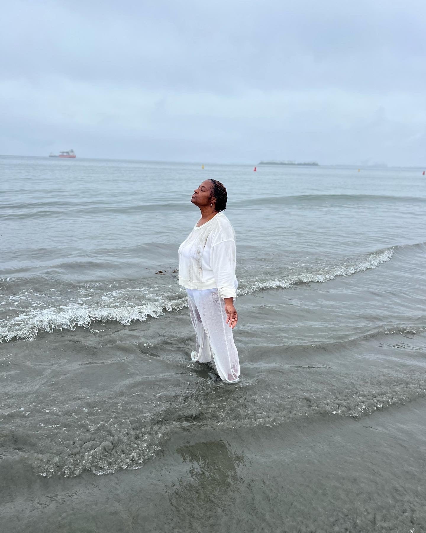 A person standing in the ocean wearing white clothing, appearing to engage in a spiritual or cleansing activity.