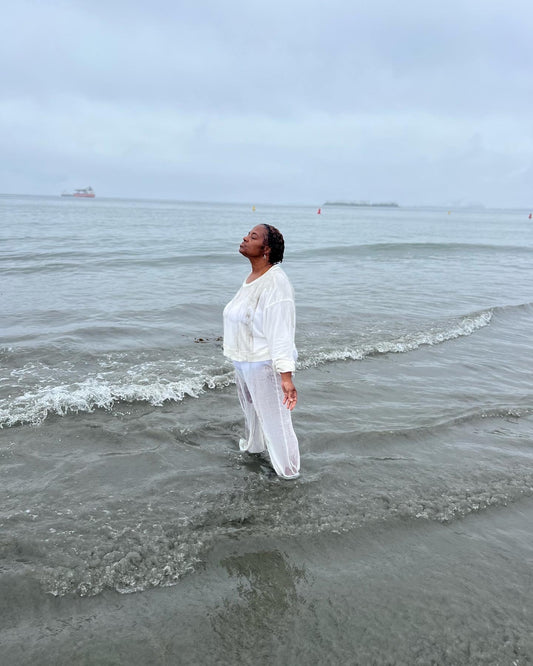 A person standing in the ocean wearing white clothing, appearing to engage in a spiritual or cleansing activity.
